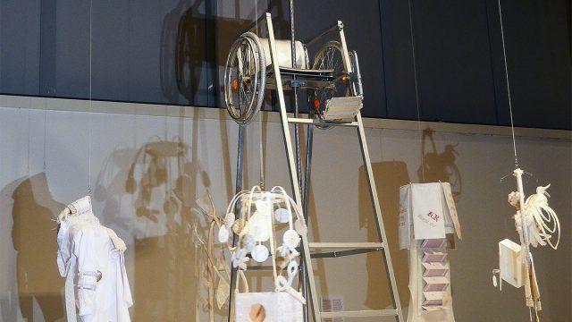 On a 3 x 3 meters pedestal covered with an enormous cloth stands a tennis judges chair. Its seat is replaced by a silver colored wheelchair. A pole rises trought the wheelchair and it is topped with a massive helicopter propeller. Hanging from the propeller blades are six textile sculptures made of fabric. There are human and animal figures, ableist texts, crutches and clothing.