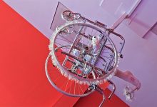 A silver colored wheelchair is making somersaults in the ceiling. The wall behind is read and the ceiling looks light purple.