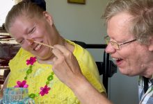 A couple with intellectual disabilities sits at a kitchen table. The man gently tickles his wife's nose with a small paintbrush. They are both laughing.