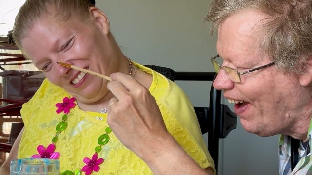 A couple with intellectual disabilities sits at a kitchen table. The man gently tickles his wife's nose with a small paintbrush. They are both laughing.