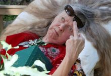 An elderly woman wearing a red beaded evening gown is lying on a pillow, long hair open. In her necklace is an owl. She stares straight into the camera and shows it her middle finger.