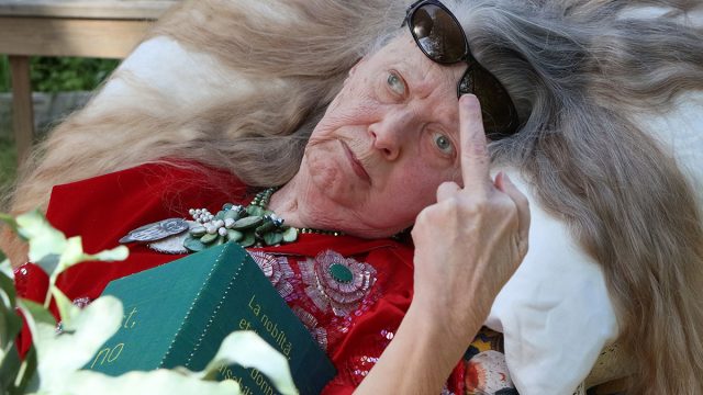 An elderly woman wearing a red beaded evening gown is lying on a pillow, long hair open. In her necklace is an owl. She stares straight into the camera and shows it her middle finger.