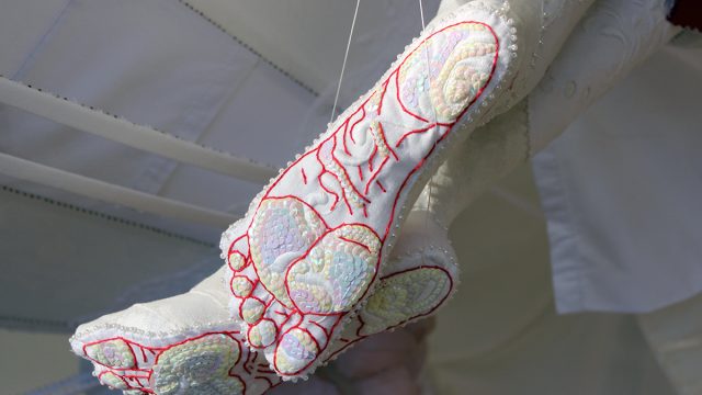 A close-up of two soles of feet from below. They are made of textile material using embroidery and are partially covered with sequins.