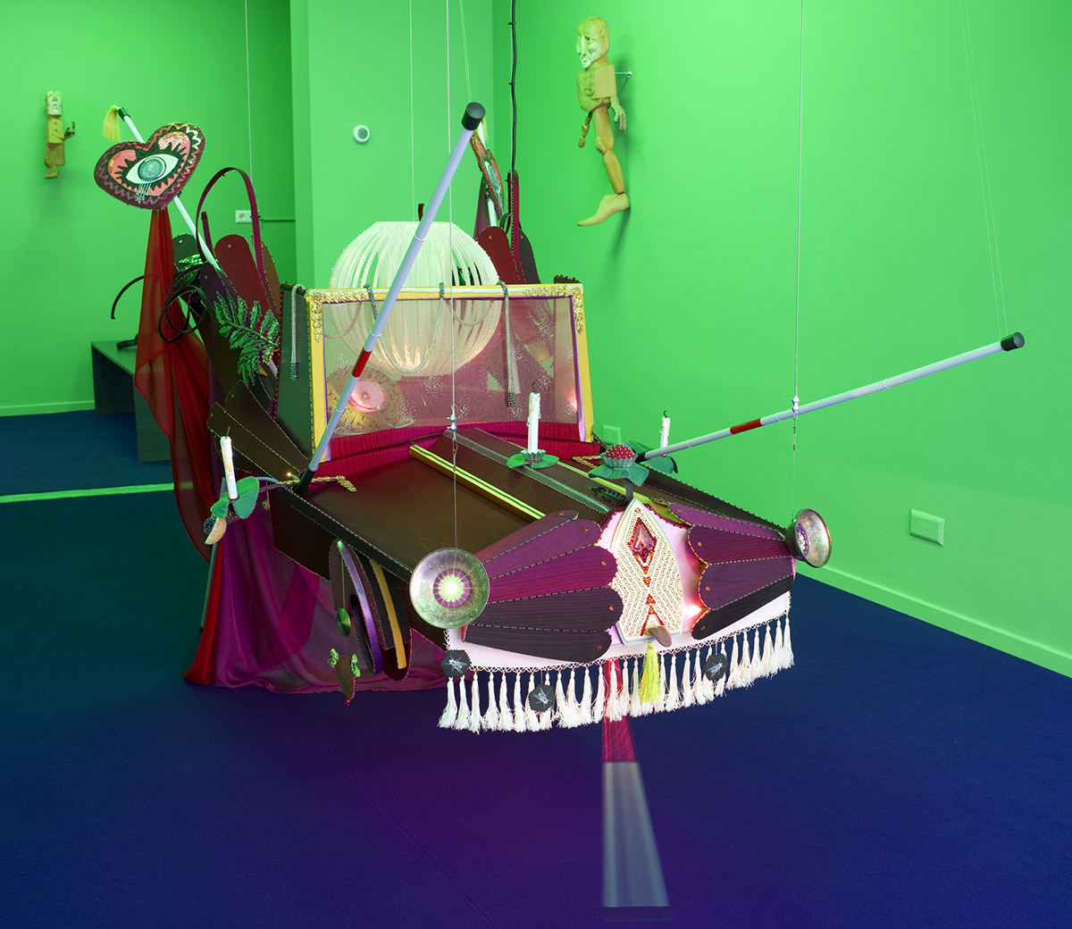 A red car sewn from textiles hangs from the gallery ceiling in a green room. White canes stick out from it, and at the front, one of the canes makes a slow side-to-side motion. The car has unlit candles in place of side mirrors, and its glittering windshield looks frosted.