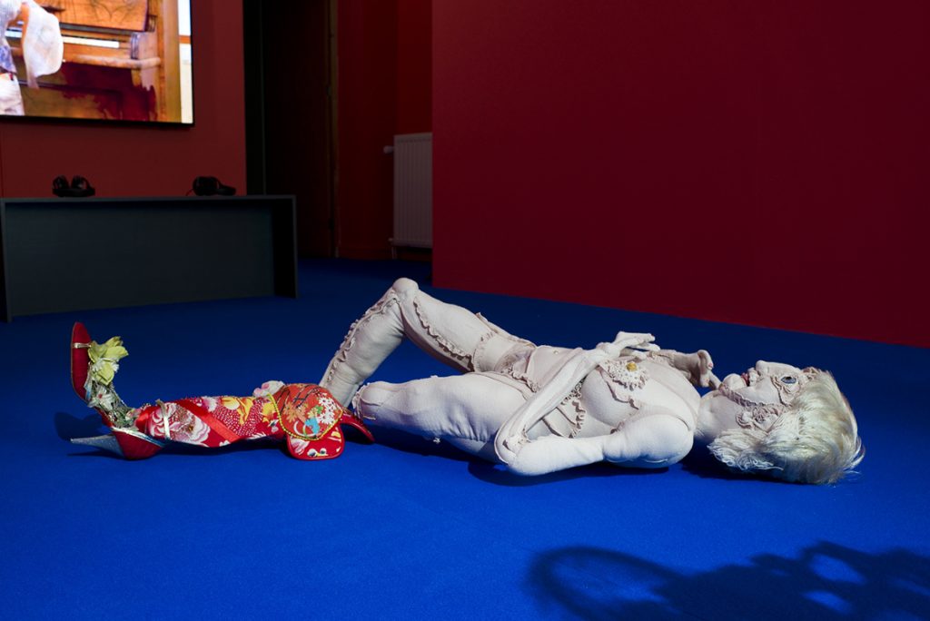 Gran-mother textile sculpture lies naked on the floor, on top of a blue carpet. Next to it is a prosthesis coverer with beading and red textiles.
