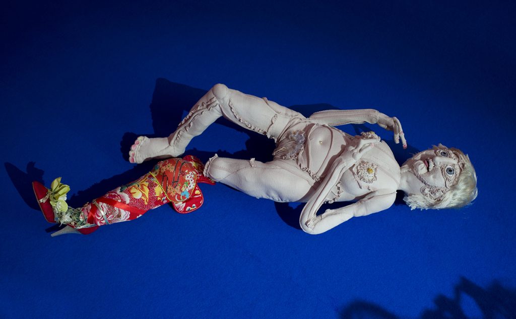 Gran-mother textile sculpture lies naked on the floor, on top of a blue carpet. Next to it is a prosthesis coverer with beading and red textiles.